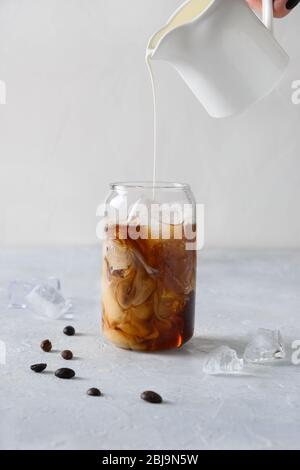 Iced latte coffee in glass with pouring milk on light background. Vertical format. Close up. Stock Photo