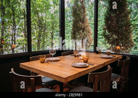 Five people dinner table with plates, knives, forks, wine glasses, glasses and napkins on wooden table with Pines and green leaf trees outside. Stock Photo
