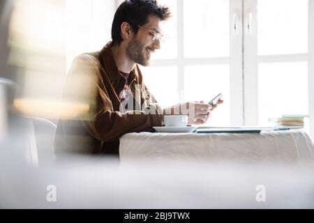 Image of handsome smiling man wearing eyeglasses using cellphone while resting in bright modern cafe Stock Photo