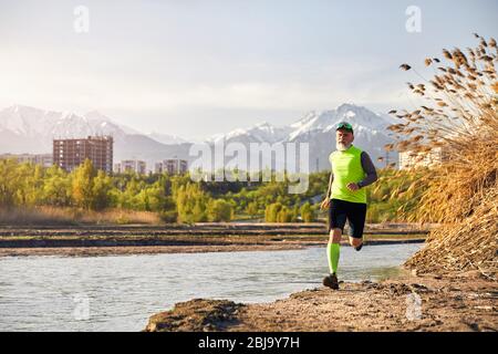 Man with grey beard running near the lake with mountain background in the morning. Healthy lifestyle concept Stock Photo