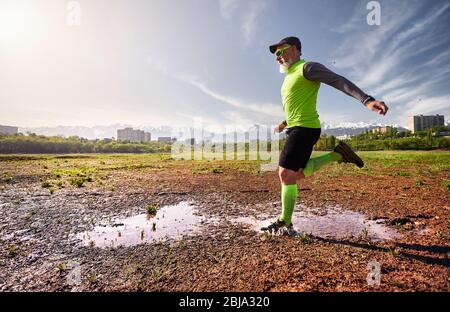 Man with grey beard running on the dirty trail with water splashes at mountain background in the morning. Healthy lifestyle concept Stock Photo