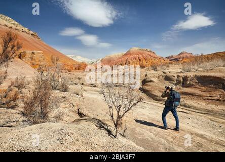 Tourist with backpack and camera taking a picture at the dusty canyon on surreal red mountains against blue sky in the desert Stock Photo
