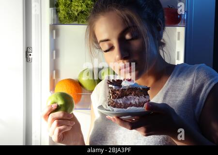 Beautiful girl eating cake and apple at night. Unhealthy food concept Stock Photo