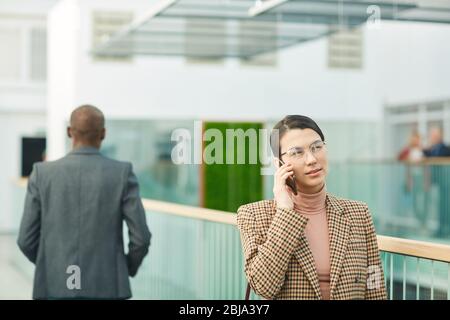 Young businesswoman in eyeglasses talking on mobile phone while walking along the office corridor Stock Photo