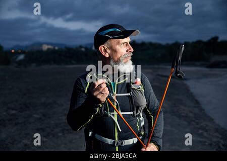 Portrait of elderly athlete man with grey beard and trekking poles at dark cloudy sky background