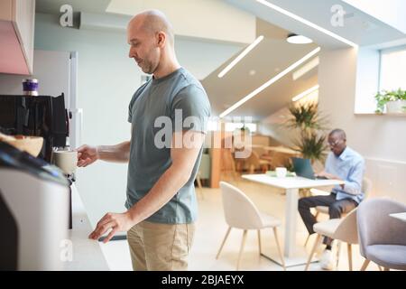 Mature man standing near the coffee machine and making coffee in the kitchen at office Stock Photo