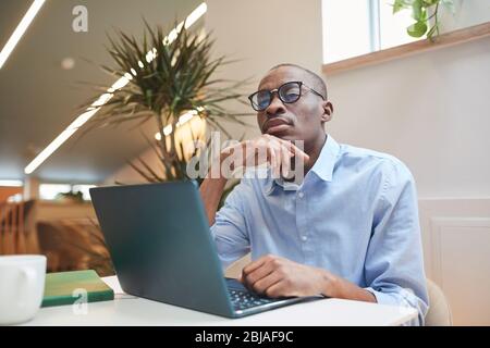 Serious African businessman in eyeglasses sitting at the table in front of laptop and working online at office Stock Photo
