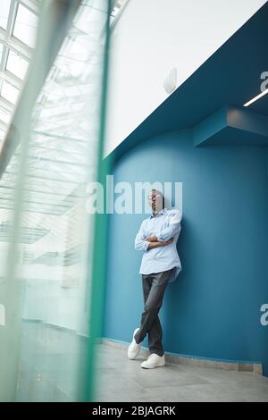 Portrait of confident African businessman leaning on the blue wall of office building and looking at camera with his arms crossed Stock Photo
