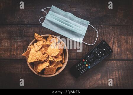 Remote control, bowl of crisps and medical mask on wooden table. Coronavirus Covid-19 quarantine stay at home concept. Stock Photo