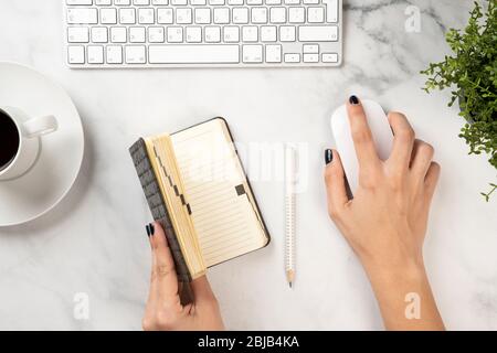 A woman holds the diary and the mouse on a white marble texture with keyboard and coffee on it Stock Photo