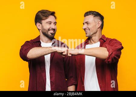 Image of two handsome men 30s in red shirts smiling and bumping their fists isolated over yellow background Stock Photo