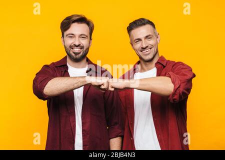 Image of two handsome men 30s in red shirts smiling and bumping their fists isolated over yellow background Stock Photo