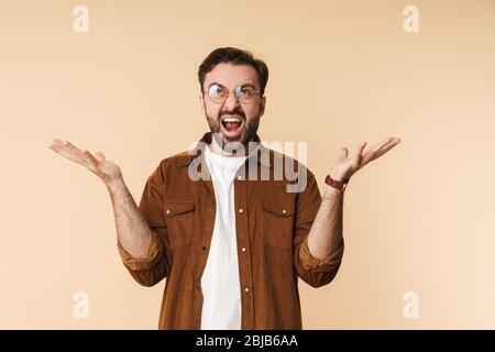 Portrait of an angry young arttractive bearded man wearing casual clothes standing isolated over beige background, screaming Stock Photo