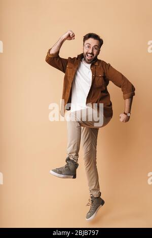 Full length portrait of a cheerful young arttractive bearded man wearing casual clothes jumping isolated over beige background, celebrating success Stock Photo