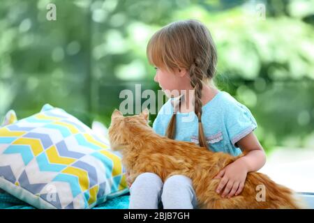 Cute little girl with red cat sitting on bed beside window Stock Photo