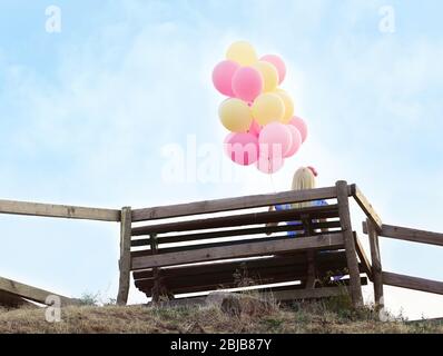 Beautiful young woman sitting on wooden bench and holding air balloons Stock Photo