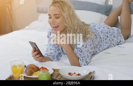 Cheerful young woman browsing smartphone and laughing lying on bed Stock Photo