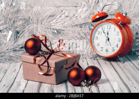 Gift box and blurred alarm clock on background on a wooden table decorated with a garland and red Christmas balls for the New Year or XMAS. Copy space Stock Photo