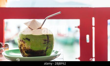 Fresh and cold whole coconut served with cut top and straw inside in beachside cafe, Koh Samui, Thailand Stock Photo