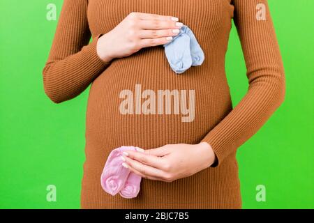 Close up of pregnant woman in brown dress holding baby pink and blue socks against her belly at green background. Is it a boy or a girl Child expectin Stock Photo