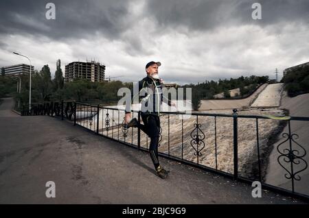 Man with grey beard in black costume and backpack running on the bridge crossing dirty river at cloudy sky background Stock Photo