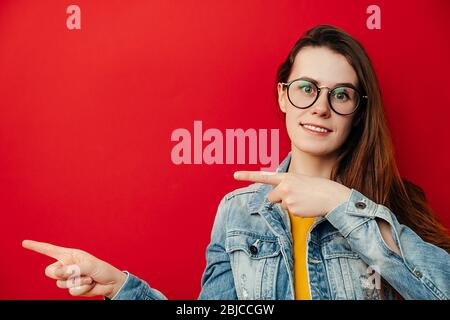 Beautiful brunette woman in eyeglasses, points with hands aside, shows copy space for your advertisement or promotional text, dressed in denim jacket, Stock Photo