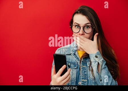 Shocked woman in eyeglasses covers mouth, feels fear, carries modern cell phone, girl read media news message online having gadget problems, dressed i Stock Photo