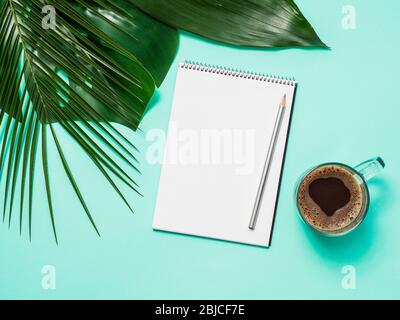 Blank paper notebook with pencil and coffee cup. Empty paper sketchbook with pincil and tropical leaves on blue background. Top view or flat lay. Copy space for text or design. Stock Photo