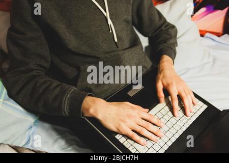Young man in casual clothes sitting on an unmade bed working remotely from home with a laptop on his knees. Closeup shot, anonymous model Stock Photo