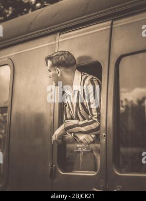 Sepia close up of isolated young 1940s man in striped blazer on board steam train, leaning & looking out of vintage railway carriage in station. Stock Photo