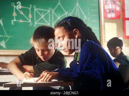 Junior school schoolgirl black African 9-11 years in classroom working at her desk with blond caucasian boy pupil geometry blackboard behind Stock Photo