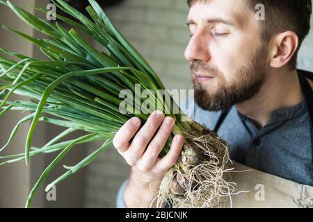 Young man in rustic apron holding and smelling a bunch of green onions. Healthy, vegetarian and organic farm food concept. Stock Photo