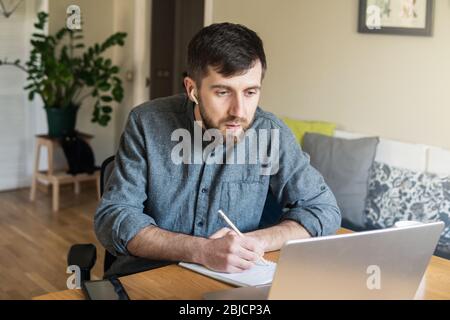 Handsome Eastern European man studying remotely at home during Corona virus quarantine lockdown, writing down during online courses Stock Photo
