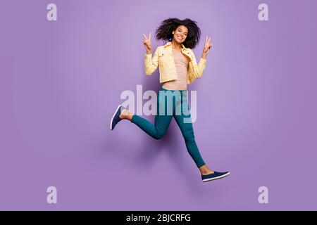 Turned full length body size photo of cheerful toothy beaming girl in pants trousers yellow shirt running junping showing v-sign isolated pastel Stock Photo