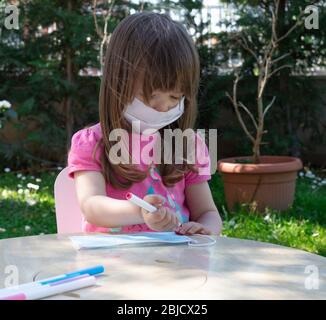 Beautiful little girl sited in the garden with her teddy bear wearing a face protection mask and coloring another one during covid-19 pandemic Stock Photo