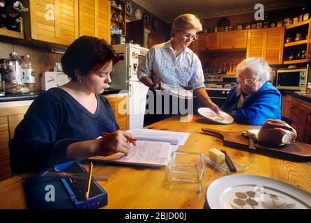 Three female generations at home living together, with elderly Grandmother, mother and granddaughter, together at meal time in their home kitchen. Mother multitasking under pressure providing meals at table for her extended family Stock Photo