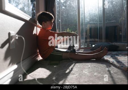 Boy sitting on the floor with ipad tablet plugged in to the wall Stock Photo