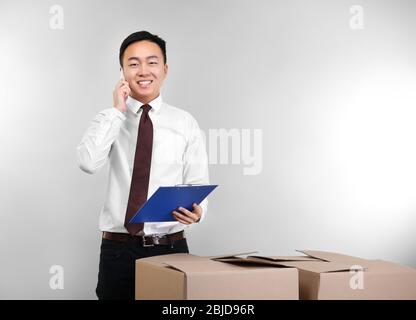 Handsome Asian man talking by mobile phone while standing near carton boxes, on light background Stock Photo