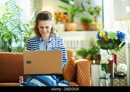happy modern woman in blue blouse and striped jacket sitting on sofa with laptop in the modern house in sunny day. Stock Photo