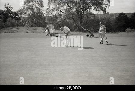 1960s, historical, outside on a wooded, inland golf course, two male golfers on a putting green, one holding the flag, the other putting, while two other golfers in the four-ball sit by the green having enjoying a cigarette, England, UK. The fashions, clubs and a golf trolley of the era can be seen. Stock Photo