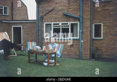 1960s, historical, a young girl sitting on a metal canvas chair in a back garden at small table with different consumer products of the era on it, having set up a pretend 'shop', England, UK. Cereals such as 'Junggle Oats' and 'Tiger Cornflakes' are displayed. Pretend play forms an essential part of a child's development, by doing and using imagination. Stock Photo