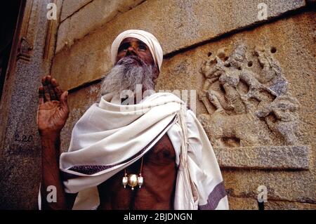 A Pujari or Hindu priest wearing traditional outfit makes the Mudra ritual hand gesture in front of an ancient shrine situated on the Hemakuta hill in the ancient village of Hampi nestled within the ruins of medieval city of Vijayanagar in the state of Karnataka in India