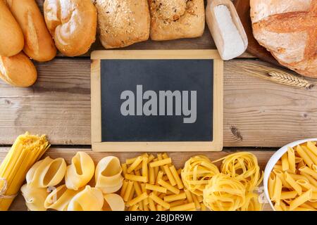 Various types of bread and pasta with  in the middle is a blackboard with copy space photographed on a wooden table in flat lay. Stock Photo