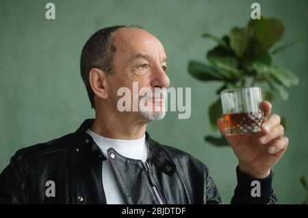 Side view of senior man holding glass of whisky or brandy and thinking about something in loft style room with light green walls and houseplants on Stock Photo