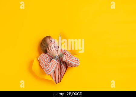 Crazy shocked, scared. Cheerful caucasian young man poses in torn yellow paper background, emotional and expressive. Breaking on, breakthrought. Concept of human emotions, facial expression, sales, ad. Stock Photo