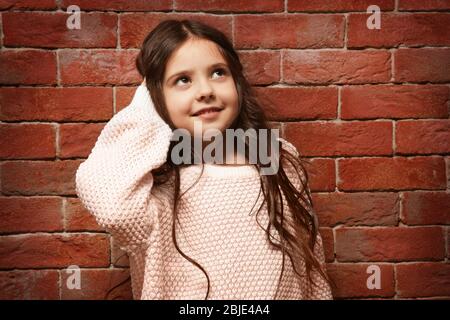 Cute little girl in warm sweater standing near brick wall Stock Photo