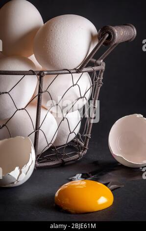 Close-up of a basket full of chicken eggs. Cracked eggshell and egg yolk in the foreground. Concept image for the proverb: don't put all your eggs in Stock Photo