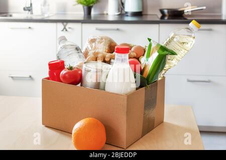 Cardboard box with various food, bottle of milk and water on the table over kitchen interior. Food donations or food safe delivery concept. Safe home Stock Photo