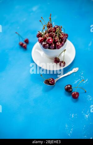 Cup of delicious ripe sweet cherries on blue background.Composition, a white ceramic Cup filled with juicy ripe red cherries, and several berries with Stock Photo