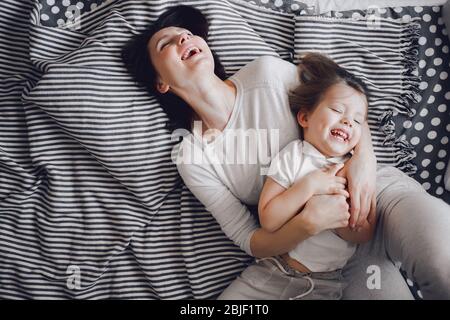 mother with 3 year old daughter hug and laugh while lying on the striped bed Stock Photo